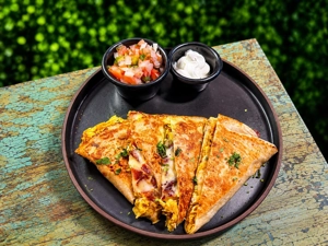 Quesadillas de pollo y tocineta con queso derretido y cilantro, acompañadas de pico de gallo y crema agria en un plato negro.