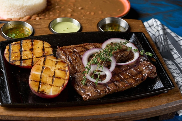 Entraña a la parrilla con aros de cebolla y rodajas de papa asada, acompañada de arroz blanco y frijoles. Cocina desconocida