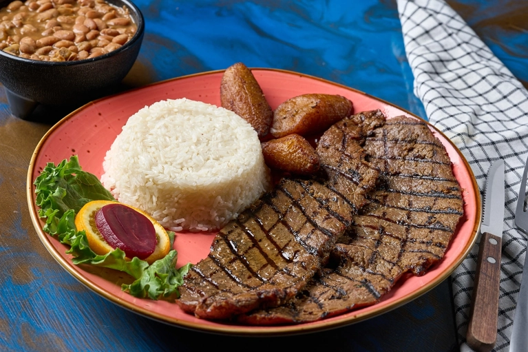 Carne asada con arroz blanco y maduros en un plato. Frijoles en un tazón al lado. Cocina desconocida en ciudad desconocida.