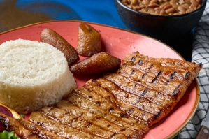 Lomo de cerdo a la parrilla con arroz blanco y maduros, acompañado de frijoles en un plato en una ciudad desconocida.