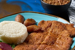 Pechuga apanada con arroz blanco y maduros visibles en el plato. Frijoles en un tazón al fondo. Cocina desconocida en ciudad