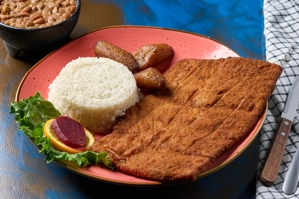 Chuleta de cerdo apanada con arroz blanco y plátanos maduros en un plato. También se ven frijoles en un tazón al fondo.