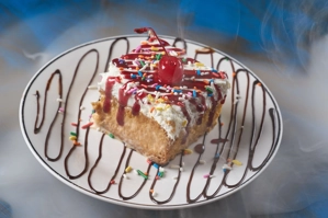 Tres leches con crema batida, cereza y chispas de colores en un plato decorado con chocolate. Cocina desconocida en ciudad