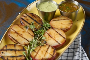 Papas cocidas a la parrilla con marcas de grill, acompañadas de brotes verdes y dos salsas en tazas metálicas. Ciudad