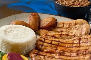 Pechuga de pollo a la parrilla con arroz blanco y plátanos maduros, acompañada de frijoles en un plato. Cocina desconocida