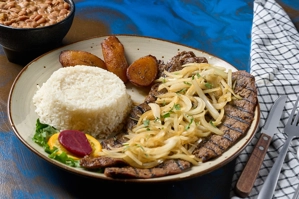 Bistec encebollado con cebolla salteada y arroz blanco, acompañado de frijoles y maduros. Cocina desconocida en ciudad