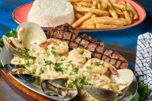 Churrasco marinado con mariscos visibles como camarones y almejas, acompañado de arroz blanco y papas fritas. Cocina
