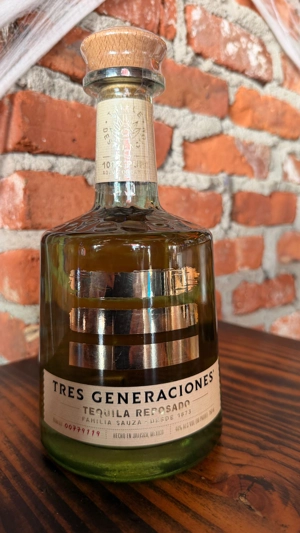 Botella de Tres Generaciones Tequila Reposado con tapa de madera sobre mesa de madera y fondo de ladrillo. Ciudad