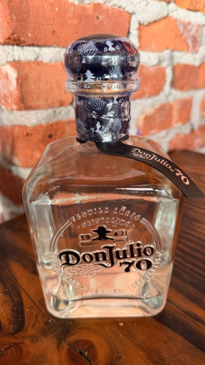 Botella de Don Julio 70 Cristalino sobre una mesa de madera, con fondo de pared de ladrillo. Tequila cristalino visible en