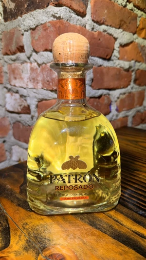 Botella de tequila Patrón Reposado sobre una mesa de madera, con fondo de pared de ladrillos. Producto mexicano.