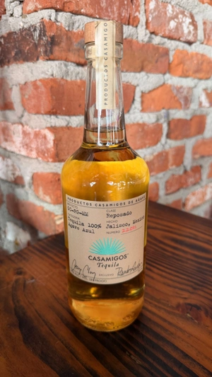 Botella de Casamigos Reposado con etiqueta visible, sobre una mesa de madera. Fondo de pared de ladrillo. Tequila de México.