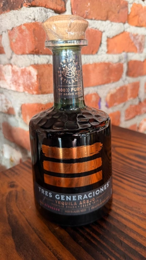 Botella de tequila Tres Generaciones Añejo con etiqueta dorada y tapa de madera sobre mesa de madera. Fondo de ladrillo