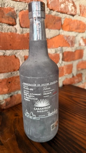 Botella de Casamigos Mezcal con etiqueta blanca y texto en español sobre una mesa de madera, fondo de ladrillo.