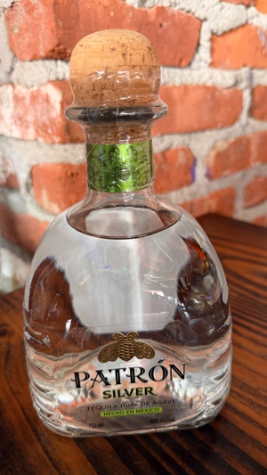 Botella de tequila Patrón Silver con tapón de corcho y etiqueta verde. Fondo de pared de ladrillo.