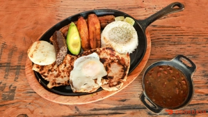 Bandeja Típica con pollo, huevo frito, arepa, aguacate y arroz en plato. Frijoles en recipiente aparte. Cocina colombiana.