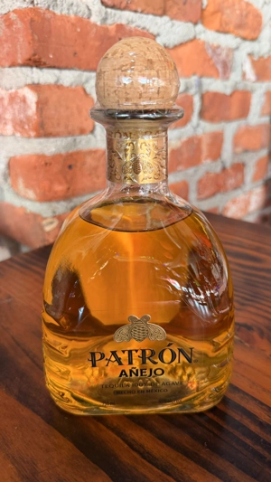 Botella de Patrón Añejo con tapa de corcho y etiqueta dorada sobre una mesa de madera. Fondo de ladrillos.