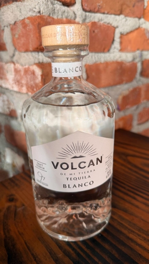 Botella de Volcan Blanco Tequila con tapa de madera y etiqueta blanca, sobre una mesa de madera, con fondo de ladrillos.