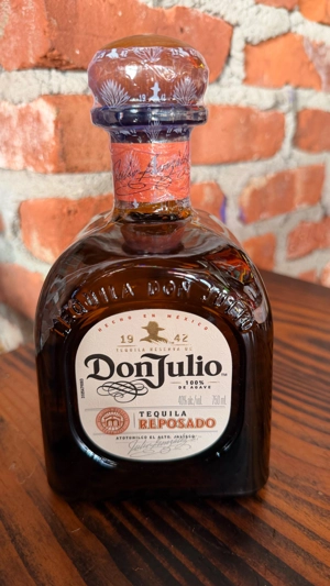 Botella de tequila Don Julio Reposado con etiqueta visible y tapa decorada. Fondo de ladrillos. Ciudad desconocida.