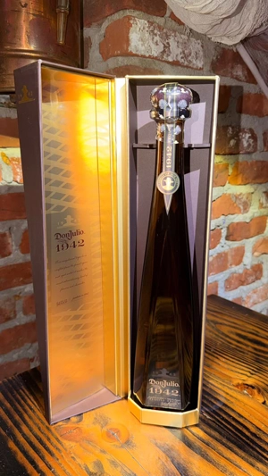 Botella de Don Julio 1942 en caja abierta sobre mesa de madera, con ladrillos de fondo. Tequila mexicano en ciudad