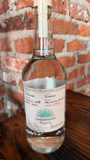 Botella de Casamigos Blanco con etiqueta visible, sobre fondo de pared de ladrillo.
