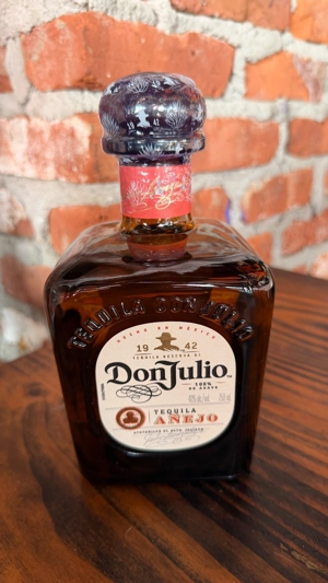 Botella de Don Julio Añejo con etiqueta blanca y tapa decorada, sobre mesa de madera. Fondo de ladrillo expuesto.