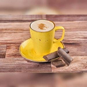 Cup of hot chocolate with frothy top and sprinkled cinnamon, next to chocolate pieces, on a wooden table. Cuisine and city