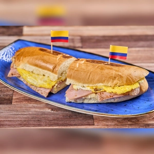 Sandwich de Jamón y Huevo with visible ham and egg, served on a blue plate with Colombian flags.