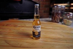Corona beer bottle on a wooden table, with a visible label and cap. No cuisine type or city is specified.