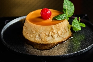Flan de café con una cereza roja y hojas de menta en un plato negro. Cocina desconocida en ciudad desconocida.