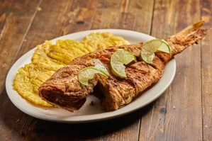 Fried red snapper with lime slices, served with tostones on a white plate. Latin cuisine in Astoria.