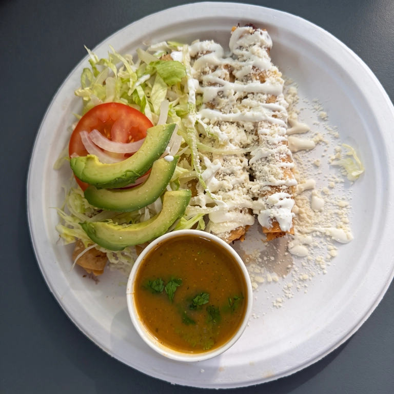 Tacos flautas with avocado slices, lettuce, and tomato on a plate in Hermiston. Served with a side of sauce.